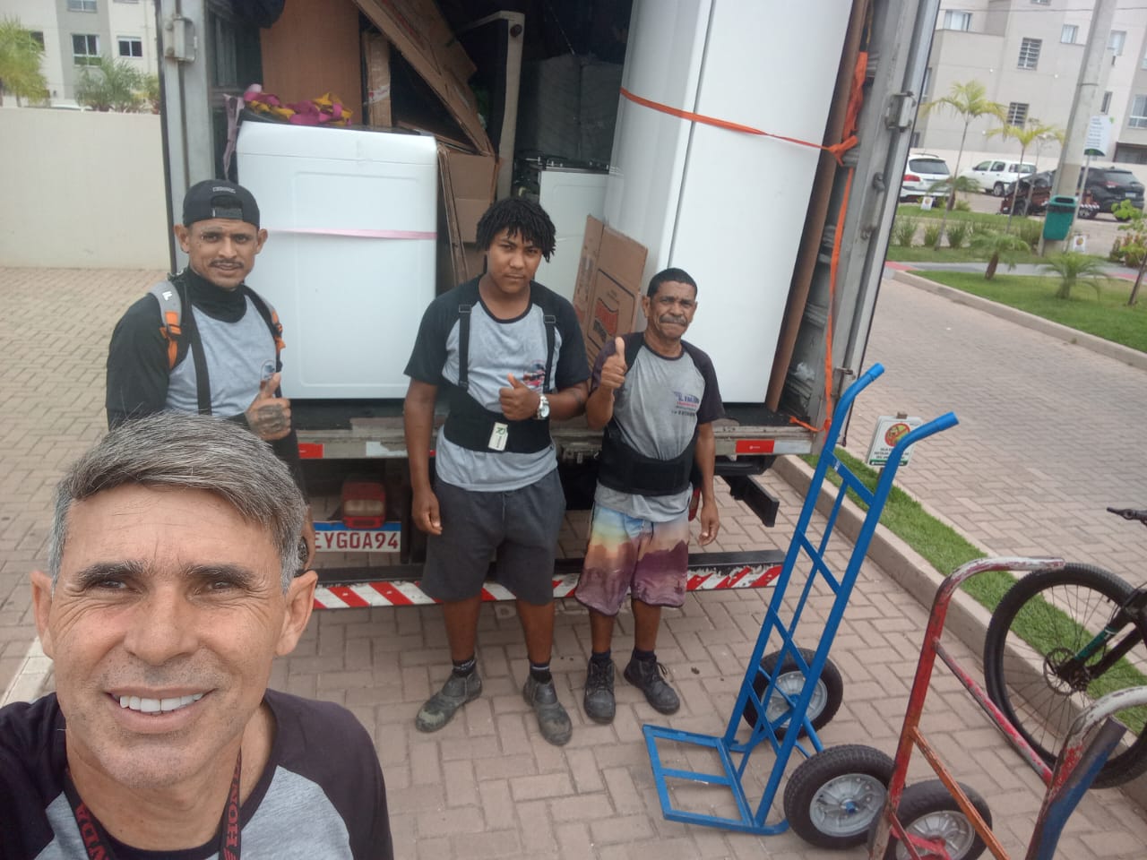Selfie do Lima com três ajudantes de uniforme cinza e cinta de proteção, fazendo joinha na frente do caminhão baú carregado